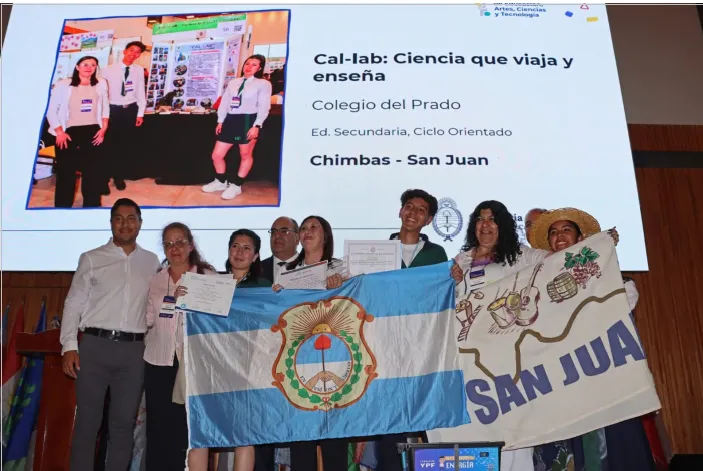 Jóvenes y adultos sonriendo en un escenario, con un cartel de premiación, celebrando el reconocimiento del proyecto CAL-LAB.