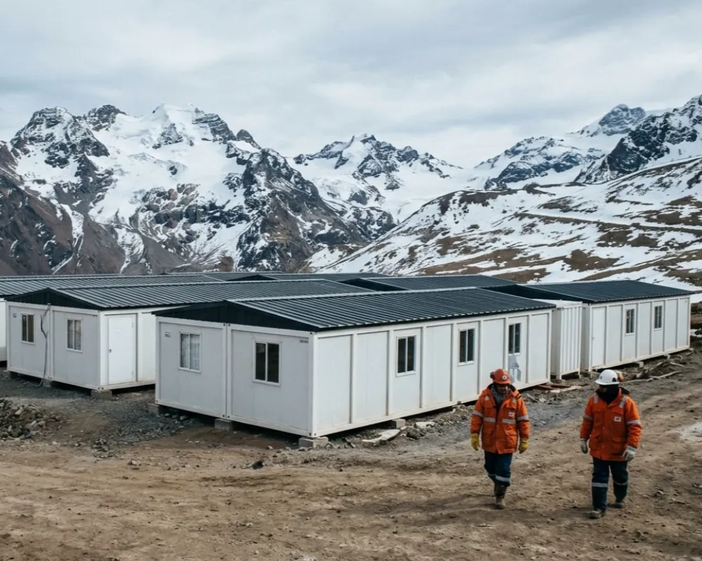 Contenido Original de ACERO Y ROCA - Prohibida su reproducción Complejo habitacional minero compuesto por módulos blancos interconectados, instalado en una plataforma rocosa a alta altitud frente a picos nevados de los Andes bajo un cielo frío. Imagen ilustrativa de Acero y Roca.