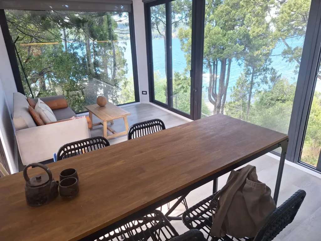 Vista desde el interior de un salón modular a través de una puerta ventana de vidrio doble, mostrando un paisaje de bosque y un lago azul de fondo.