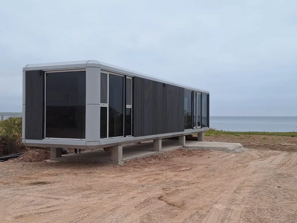 Casa modular de diseño contemporáneo, color gris con amplios ventanales, elevada sobre pilares de hormigón en un entorno natural despejado