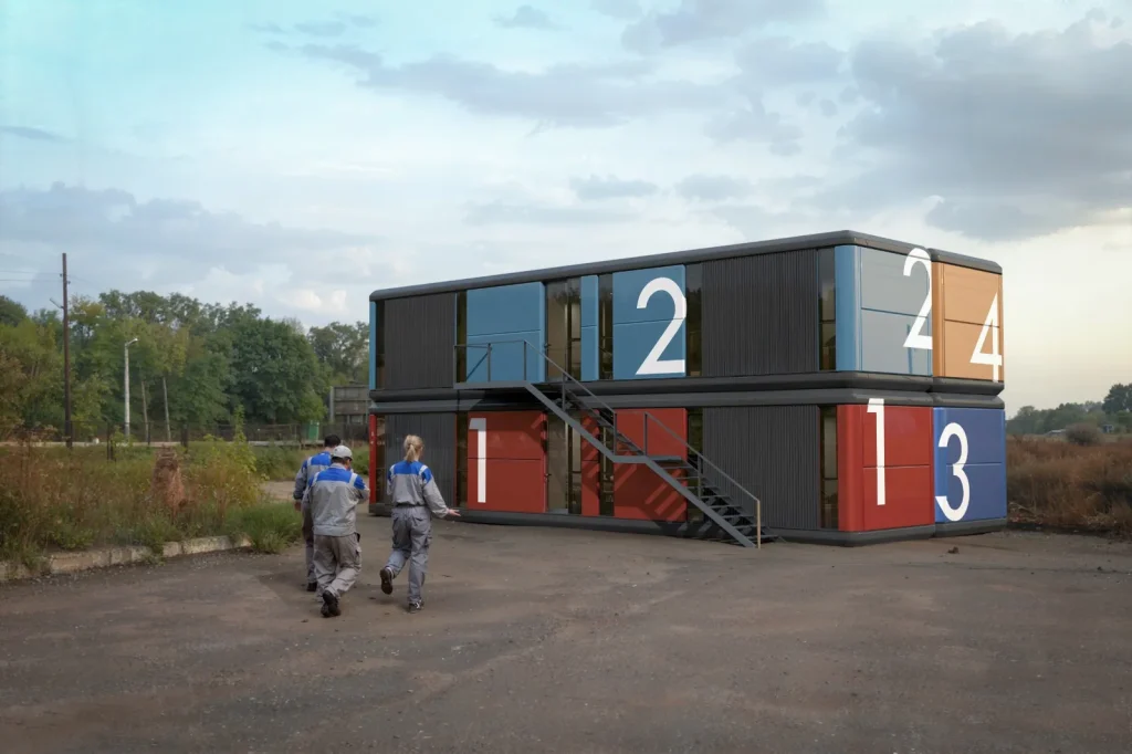 Vista exterior de un edificio modular de dos plantas compuesto por cuatro contenedores habitacionales grises de diseño moderno, con grandes ventanales y una escalera metálica lateral que conecta ambos niveles en un entorno despejado.