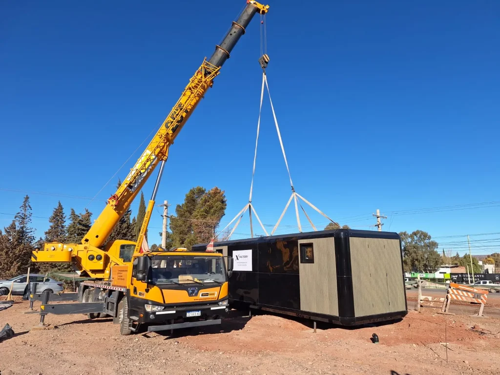 Una grúa hidráulica levantando en el aire una estructura modular gris para colocarla sobre un terreno inclinado rodeado de pinos.