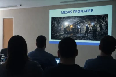 Participantes de espaldas en una mesa técnica del ProNaPre observan una presentación proyectada con el título “Mesas PRONAPRE” y una imagen de minería subterránea con maquinaria pesada y operarios en operación.