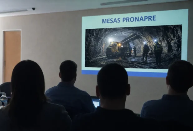 Participantes de espaldas en una mesa técnica del ProNaPre observan una presentación proyectada con el título “Mesas PRONAPRE” y una imagen de minería subterránea con maquinaria pesada y operarios en operación.