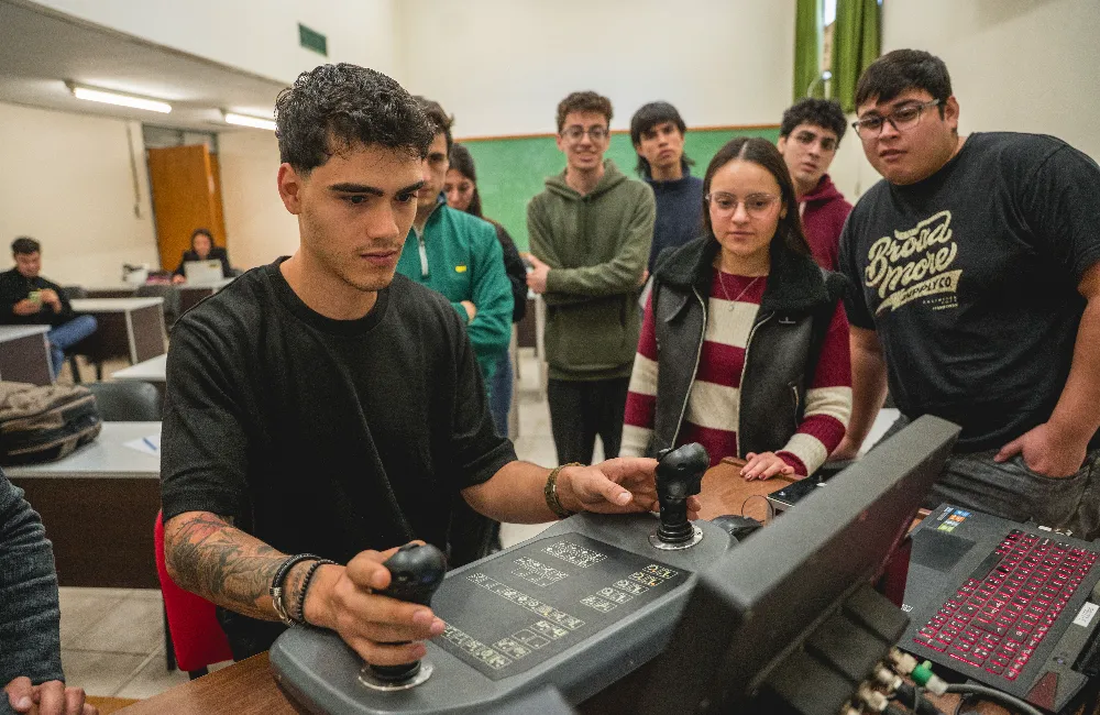 Fotografía de un estudiante de ingeniería operando una estación de simulación de escritorio con joysticks de control dual mientras sus compañeros observan el procedimiento.