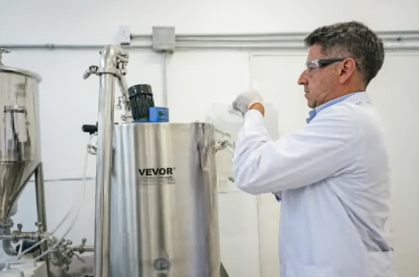 técnico con bata blanca y gafas de seguridad operando un gran reactor industrial de acero inoxidable.  