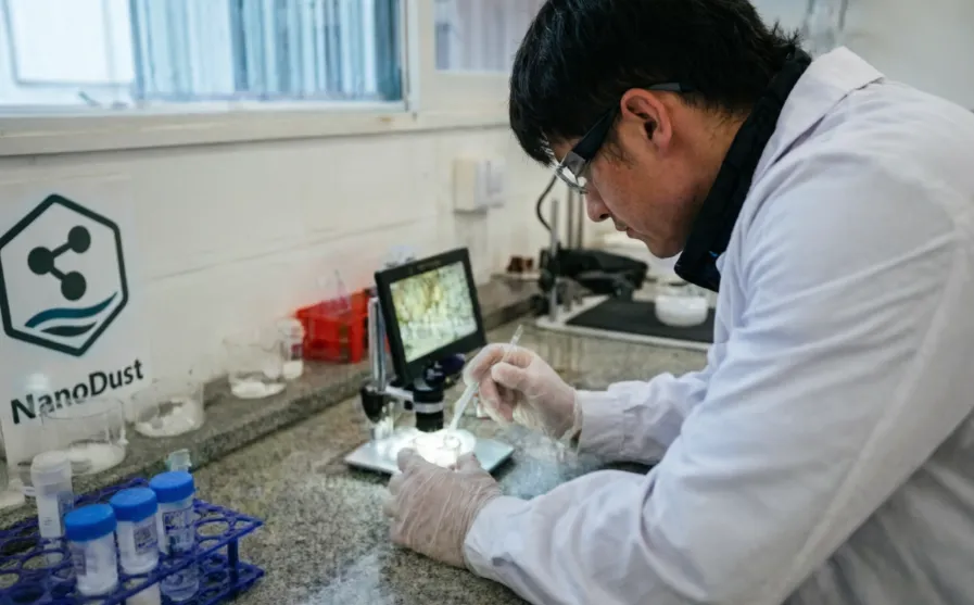 investigador con bata blanca y gafas de seguridad manipulando muestras con micropipeta bajo un estereomicroscopio en un laboratorio de nanotecnología. 