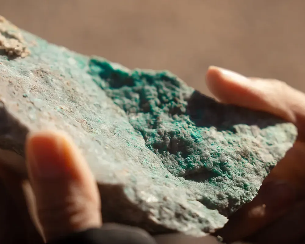Manos en guantes de cuero sosteniendo mineral de cobre crudo ante plano holográfico flotante de satélite.