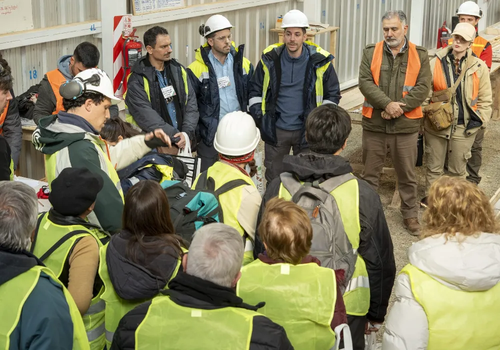 Un plano general interior que muestra a un grupo de personas reunidas. En el fondo, varios profesionales con cascos de seguridad blancos y chaquetas o chalecos de alta visibilidad (amarillo y azul) escuchan a un orador con un casco blanco que gesticula hacia la izquierda. En primer plano, varias personas vestidas con chalecos de alta visibilidad amarillos y mochilas están de pie, vistas desde atrás, observando la presentación. El entorno parece ser un espacio de trabajo técnico o de exposición, con estanterías y un extintor visible.