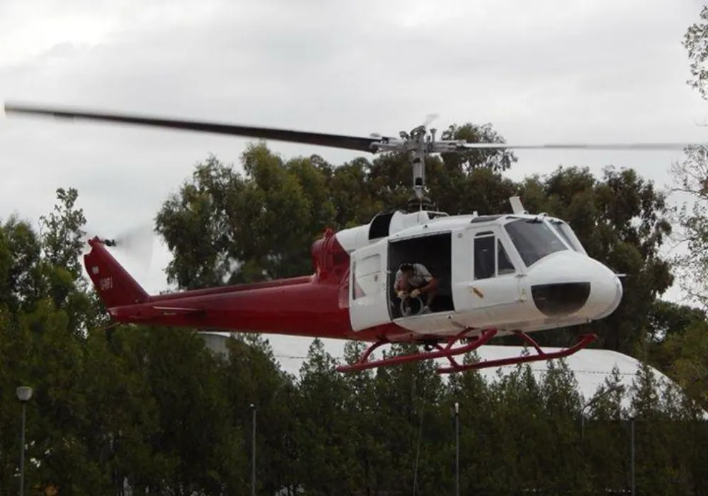 Helicóptero blanco y rojo en vuelo con un tripulante en la puerta lateral abierta.