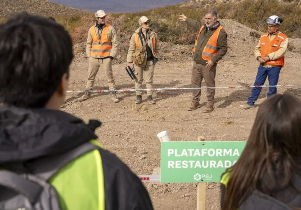 Un plano medio exterior en un paisaje árido de montaña. En primer plano, las cabezas y hombros de dos observadores con chalecos de alta visibilidad amarillos (uno con una mochila) están de espaldas. Delante de ellos, un cartel verde brillante dice "PLATAFORMA RESTAURADA" en letras blancas grandes, con el logo "PSJ" debajo. Más allá del cartel, tres hombres con uniformes de seguridad (chalecos naranjas y cascos, uno con una gorra de béisbol) están de pie, uno de ellos gesticula hacia las montañas secas al fondo. Una cinta de advertencia roja y blanca delimita la zona.