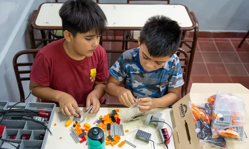 Dos niños observan y clasifican piezas de un kit de robótica sobre una mesa. Frente a ellos hay una caja organizadora con componentes plásticos y mecánicos que utilizarán para construir un proyecto tecnológico.