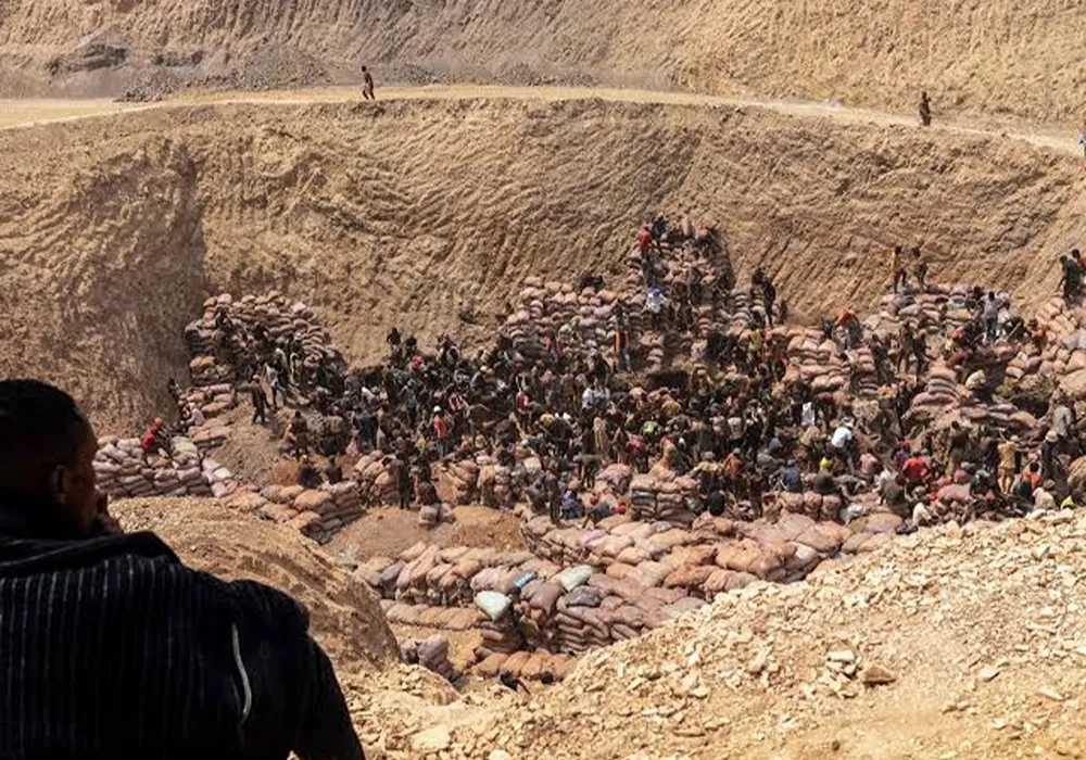 Vista desde una colina de una enorme explotación minera artesanal, mostrando a cientos de personas trabajando con palas y picos en un paisaje erosionado, con colinas áridas y cielo despejado de fondo