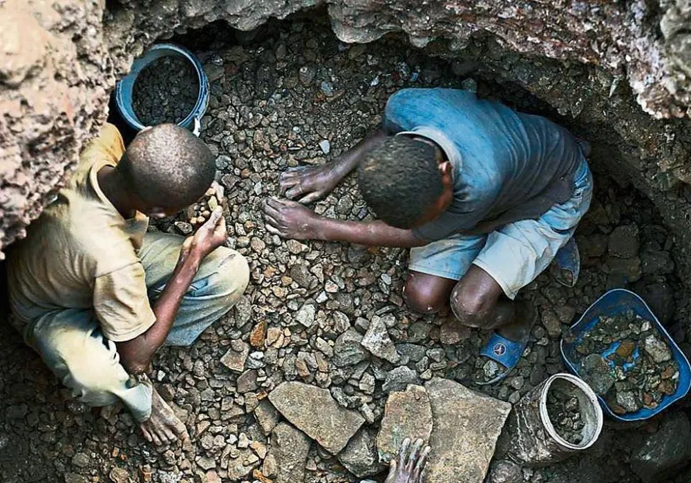 Dos mineros congoleños arrodillados dentro de una zanja estrecha, clasificando piedras de cobalto a mano, con cubetas y herramientas rudimentarias a su alrededor.