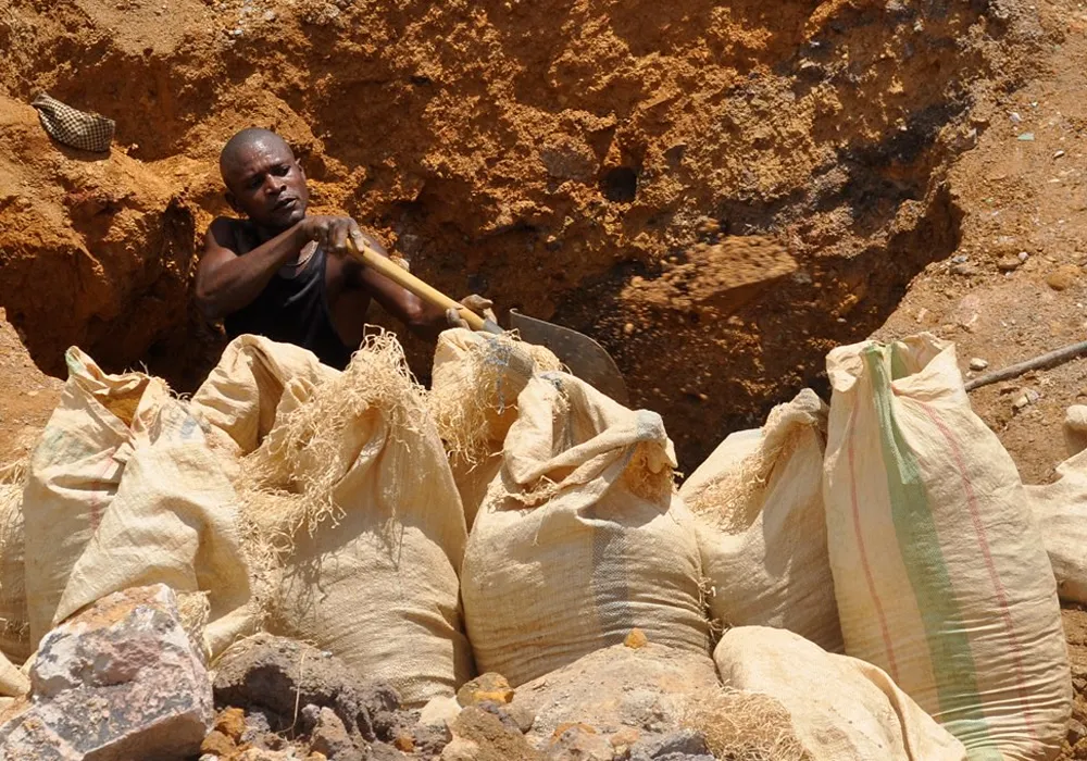 Primer plano de un minero congoleño sin equipo de seguridad, paleando tierra en una mina a cielo abierto, con varios sacos de arpillera llenos de mineral en primer plano.