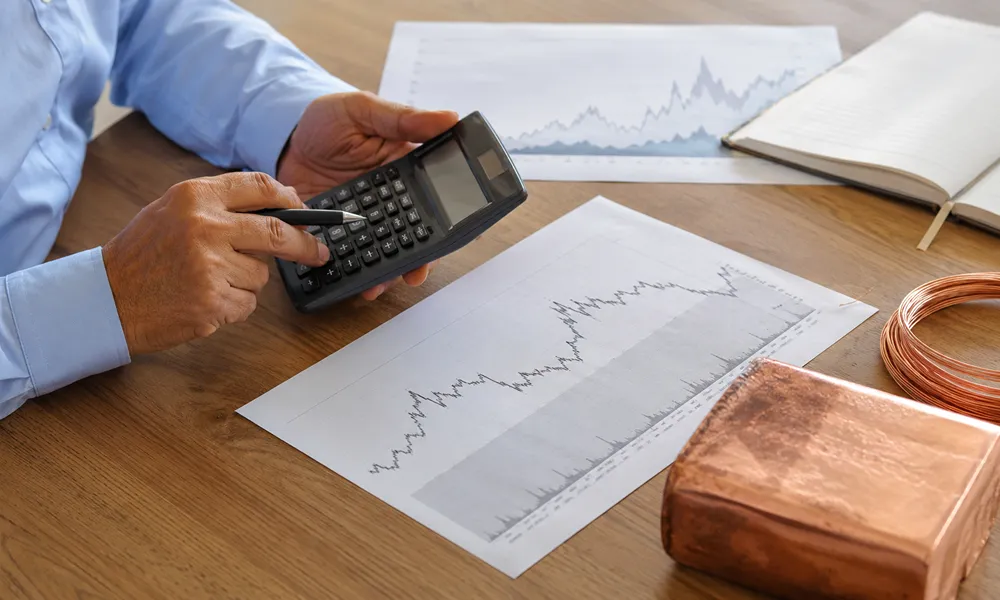 Un inversor utiliza una calculadora y sostiene un bolígrafo sobre gráficos de cotizaciones financieras impresos, con una bobina de alambre de cobre y un bloque de metal de cobre a su lado en una mesa de madera.