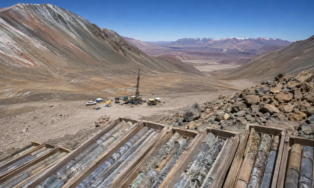 Una vista panorámica de un gran campamento de exploración minera en una zona montañosa árida de los Andes, con picos nevados a lo lejos. En primer plano, cajas de madera llenas de núcleos de perforación de roca extraídos están alineadas.