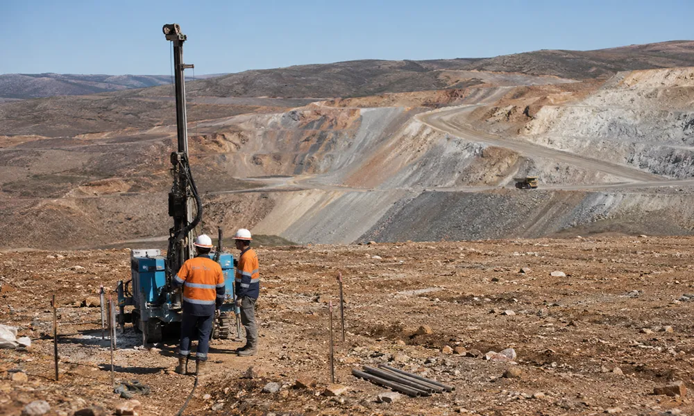 Dos trabajadores con ropa de trabajo minera (camisas naranjas y cascos blancos) están de pie junto a una pequeña máquina perforadora de núcleo en un sitio de proyecto a gran escala en Los Azules. Al fondo se ven grandes bancos de excavación de tajo abierto y un camión minero.