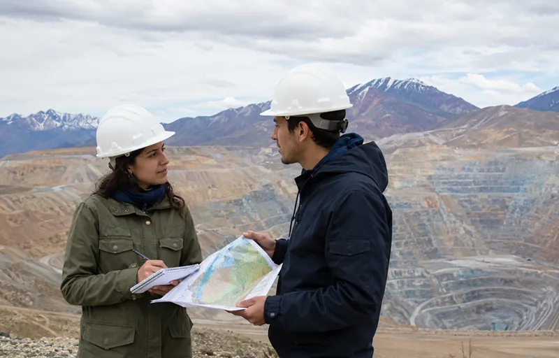 Dos jóvenes con cascos observan mapas y toman notas en un mirador natural frente a una mina a cielo abierto, con terrazas del yacimiento y cordillera al fondo.