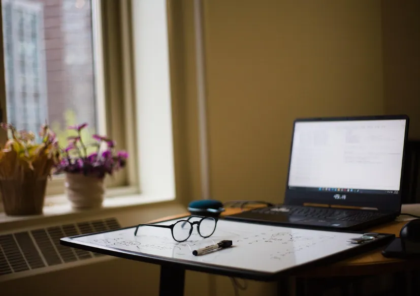 Computadora sobre una mesa con anteojos y una pizarra.
