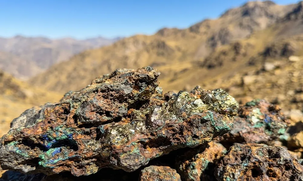Primer plano de mineral de cobre con vetas rojizas y doradas de calcopirita ante el paisaje cordillerano desenfocado bajo el sol de alta montaña.
