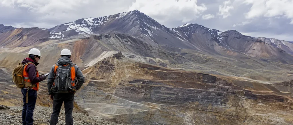 Dos técnicos observan una explotación minera a cielo abierto de gran escala emplazada en un ambiente montañoso, con bancos de excavación visibles sobre el relieve.