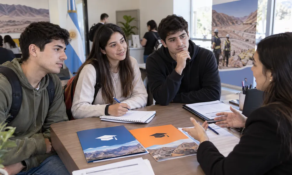 Estudiantes universitarios reciben orientación sobre un programa de becas en una oficina pública de San Juan, con documentación académica y referencias visuales a la actividad minera.