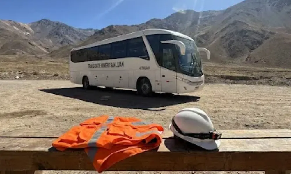 Colectivo de transporte de pasajeros de larga distancia homologado para personal minero estacionado en la cordillera de San Juan con casco y chaleco de alta visibilidad en primer plano.